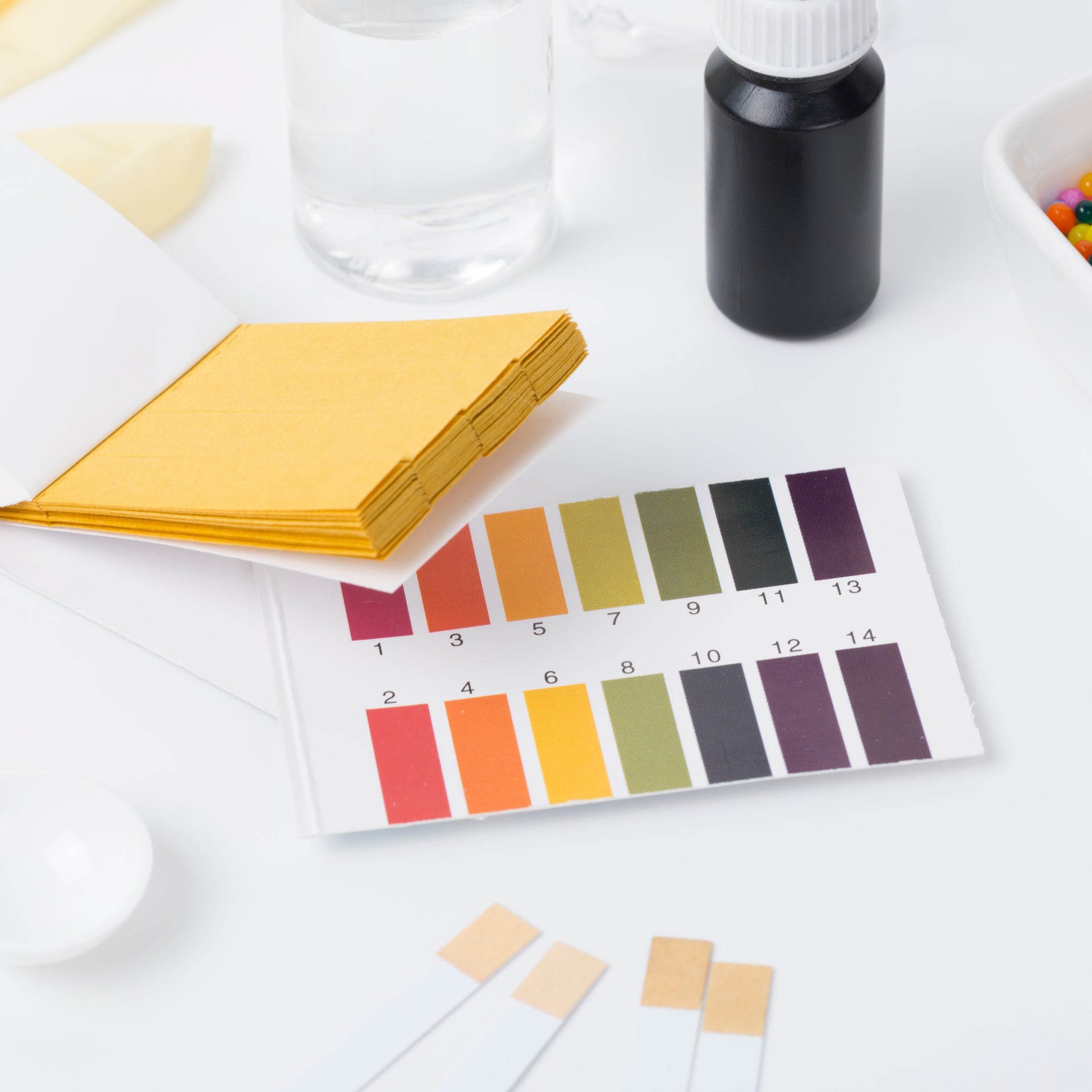 <On a white lab worktop, a colour pH chart of indicators from red and pH 1 via orange to pH4, yellow pH 6, green pH 7, and violet pH 10 to 14, next to two sample bottles and test strips. Credit> AAdobeStock_216414398