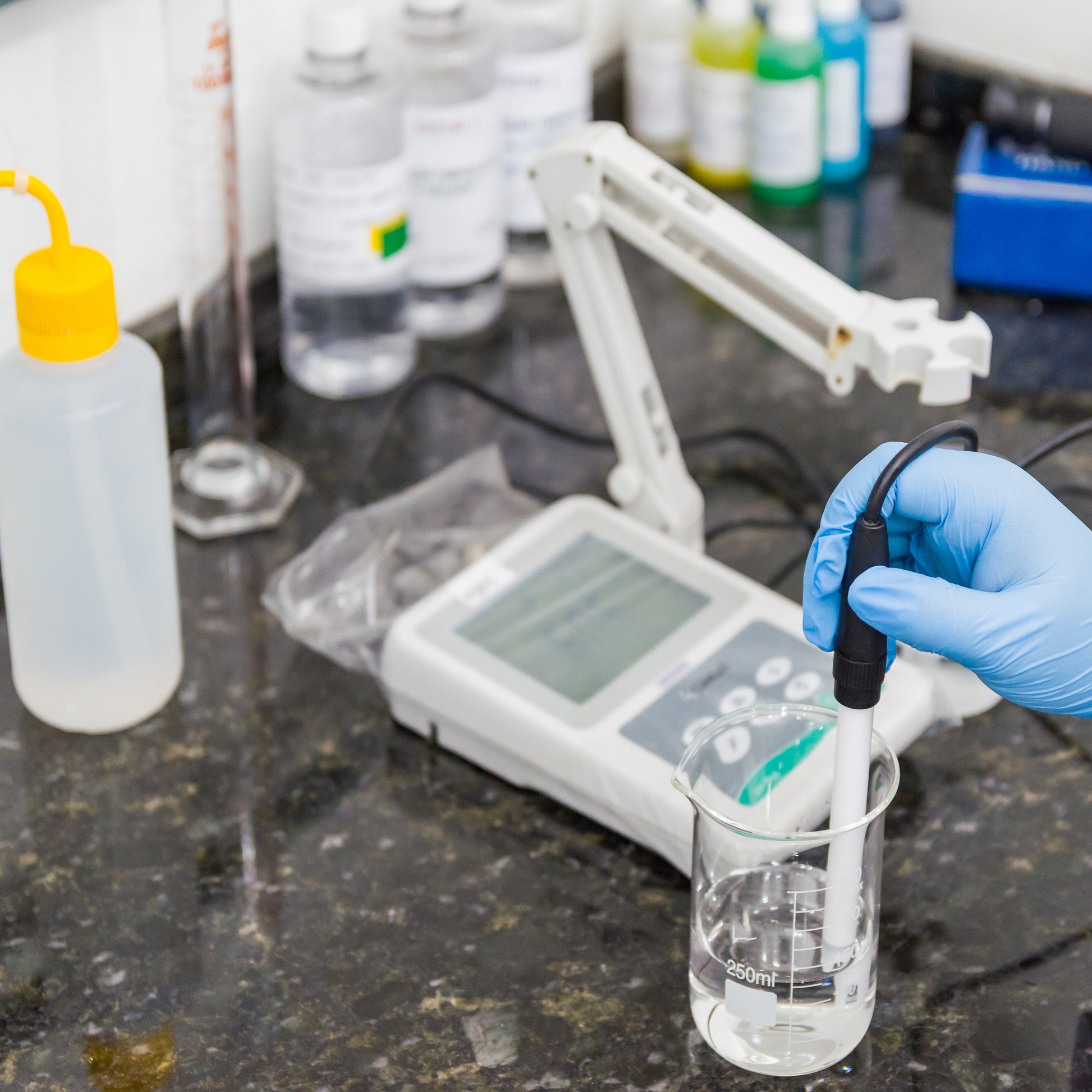 <Digital white pH meter on a black stone effect lab worktop with a range of buffers, and an analyst hand wearing blue gloves holding the white probe in a glass beaker of colourless liquid. Credit> AdobeStock_296409151